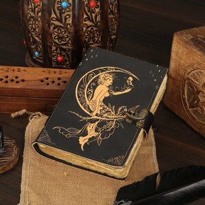 May include: A black leather journal with a gold embossed design of a mermaid sitting on a crescent moon. The journal has a clasp closure and is decorated with stars.