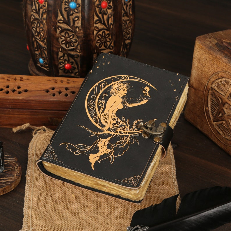 May include: A black leather journal with a gold embossed design of a mermaid sitting on a crescent moon. The journal has a clasp closure and is decorated with stars.