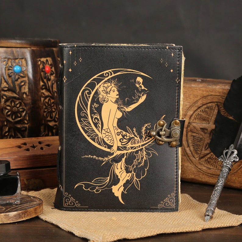 May include: A black leather journal with a gold embossed design of a mermaid sitting on a crescent moon. The journal has a metal clasp closure.