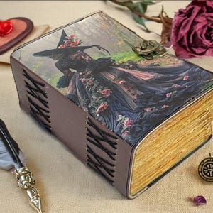 Puede incluir: Una caja decorativa con forma de libro con una ilustraci&oacute;n de bruja en la portada. La caja tiene una cubierta de cuero marr&oacute;n con costuras negras y un cierre de metal. Las p&aacute;ginas son de color dorado. Una pluma, un objeto en forma de coraz&oacute;n y una rosa est&aacute;n cerca.