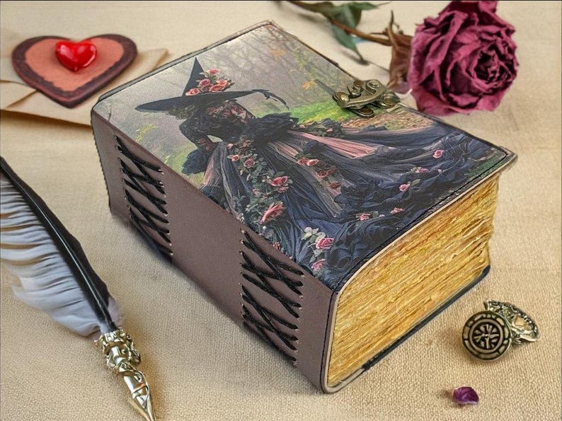 Puede incluir: Una caja decorativa con forma de libro con una ilustraci&oacute;n de bruja en la portada. La caja tiene una cubierta de cuero marr&oacute;n con costuras negras y un cierre de metal. Las p&aacute;ginas son de color dorado. Una pluma, un objeto en forma de coraz&oacute;n y una rosa est&aacute;n cerca.