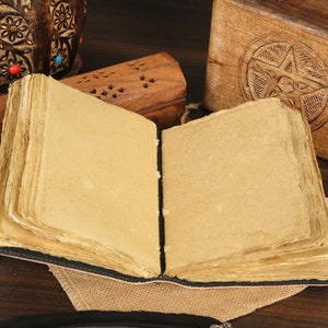 May include: An open, blank journal with a brown leather cover and worn, aged paper inside. The journal is surrounded by wooden boxes and a small inkwell, suggesting a writing or journaling setting.