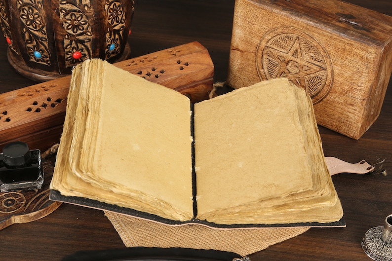 May include: An open, blank journal with a brown leather cover and worn, aged paper inside. The journal is surrounded by wooden boxes and a small inkwell, suggesting a writing or journaling setting.