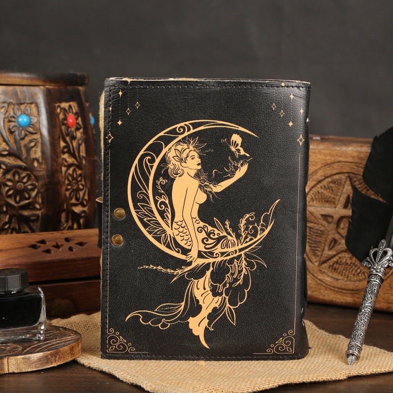 May include: Black leather journal featuring a gold design of a mermaid sitting on a crescent moon. The mermaid has long flowing hair and is holding a butterfly.