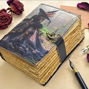 Puede incluir: Un diario de estilo vintage con una ilustraci&oacute;n g&oacute;tica en la portada de una mujer con un vestido oscuro. El diario tiene p&aacute;ginas envejecidas y amarillentas, una correa de cuero negro y un cierre de metal. Dos plumas estilogr&aacute;ficas y rosas secas est&aacute;n cerca.