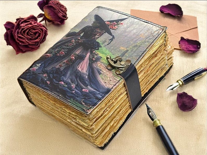 Puede incluir: Un diario de estilo vintage con una ilustraci&oacute;n g&oacute;tica en la portada de una mujer con un vestido oscuro. El diario tiene p&aacute;ginas envejecidas y amarillentas, una correa de cuero negro y un cierre de metal. Dos plumas estilogr&aacute;ficas y rosas secas est&aacute;n cerca.
