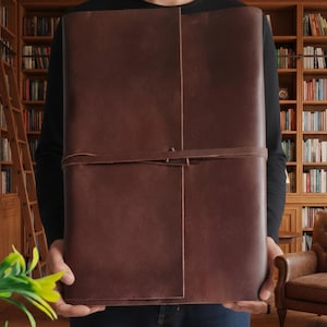 May include: A large, brown leather journal with a wrap-around strap closure. The journal is held in front of a person, with a library background visible. The leather appears to be a rich, dark brown colour.