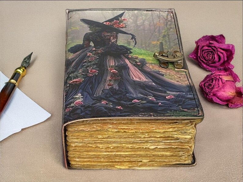 Puede incluir: Un diario encuadernado en cuero con una ilustraci&oacute;n de bruja en la portada, con una mujer con un vestido y sombrero negros con rosas rosas. El diario tiene p&aacute;ginas amarillentas envejecidas y un cierre de metal. Un bol&iacute;grafo y papel est&aacute;n a la izquierda.