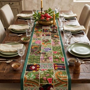 Op de afbeelding: Een rechthoekige tafelloper met een patchworkontwerp van groene stoffen vierkanten met gedetailleerd, meerkleurig bloemen- en paisley-borduurwerk. De loper ligt op een houten eettafel met borden, servetten, glazen en een schaal met granaatappels.