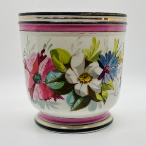 Puede incluir: Una taza de cerámica blanca con una banda rosa y un borde dorado alrededor del borde. La taza está decorada con flores pintadas a mano en tonos de rosa, blanco, azul y verde.