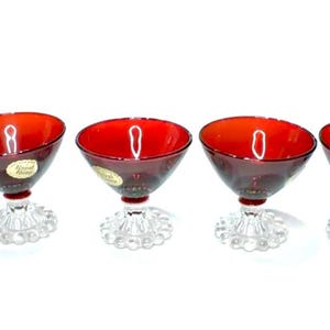 May include: Five ruby red cocktail glasses with clear, textured stems. The glasses have a unique, angled bowl design and are arranged in a row against a white background. Each glass has a small, gold sticker.