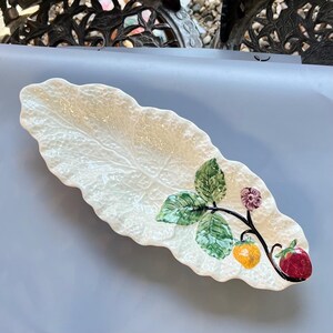 Op de afbeelding: Crèmekleurige, bladvormige keramische serveerschaal. De schaal is versierd met een beschilderde tak met groene bladeren, een gele vrucht, een paarse bloem en een rode aardbei. De schaal heeft een getextureerd oppervlak.