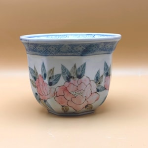 May include: A white ceramic cup with a floral design. The cup features pink flowers with green leaves and a blue geometric pattern around the rim. The cup has a slightly flared shape.
