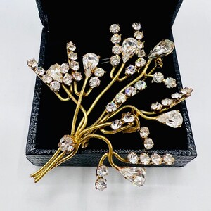 Puede incluir: Un broche de tono dorado con un diseño floral con brillantes de cristal transparentes. El broche tiene la forma de un ramo de flores con tallos y hojas delicados.