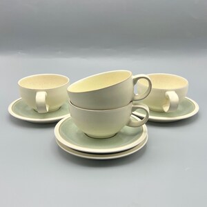 May include: Cream-colored teacups and saucers. The cups have a rounded shape with a handle. The saucers are light green with a cream-colored rim. The set is arranged on a light gray surface.