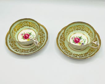 May include: Two vintage teacups and saucers. The cups and saucers are light green with gold filigree trim. Each cup features a floral design with a pink rose and other colorful flowers. The handles are white with gold accents.