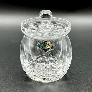 Könnte beinhalten: Ein klarer Kristallbehälter mit Deckel. Der Behälter hat einen runden Körper und einen Deckel mit einem kleinen Knopf. Der Kristall ist mit einem Diamantenmuster geschnitten. Ein blaues und goldenes Etikett ist am Behälter angebracht.