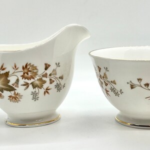 Peut inclure: Un ensemble de pot à crème et de sucrier blanc avec un motif floral brun et vert. L'ensemble est orné d'une bordure dorée.