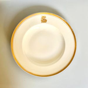 May include: Cream-colored plate with a gold rim and a gold emblem at the top. The plate has a wide rim and a shallow center. The gold rim adds a touch of elegance to the plate's design. This plate is suitable for serving food.