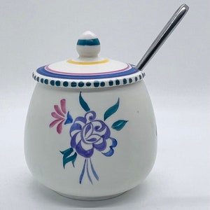 Può includere: Zuccheriera in ceramica bianca con un motivo floreale in tonalità di blu, verde e rosa. Il coperchio ha una fascia gialla e una striscia verde e blu. È inclusa una cucchiaino d'argento.