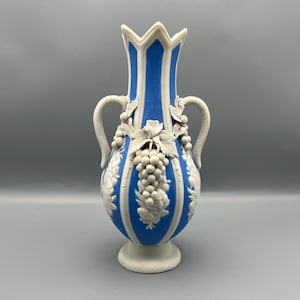 Puede incluir: Jarrón de cerámica azul y blanco con un diseño en relieve detallado de uvas y hojas. El jarrón tiene una parte superior acampanada, dos asas y una base de pedestal. El jarrón mide aproximadamente 25 cm de alto.