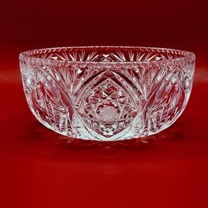 May include: A clear, round crystal bowl featuring intricate cut glass patterns, including a prominent central starburst motif surrounded by fan-shaped cuts and a beaded rim. The bowl has a reflective surface and deep, geometric facets throughout.