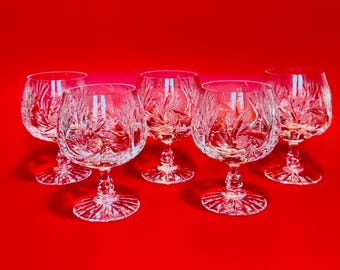 May include: Five clear crystal brandy glasses with detailed starburst and leaf patterns. The glasses have a rounded bowl, a slender stem, and a detailed base. The background is a solid red.