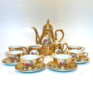May include: A gold-colored tea set with a teapot, creamer, sugar bowl, and multiple teacups and saucers. Each piece features a painted scene and gold trim. The set is arranged on a white surface.