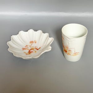 Op de afbeelding: Een witte keramische kom en beker set. De kom heeft een geschulpte rand en een bloemenontwerp in tinten oranje en rood. De beker is cilindrisch met een vergelijkbaar bloemenpatroon.