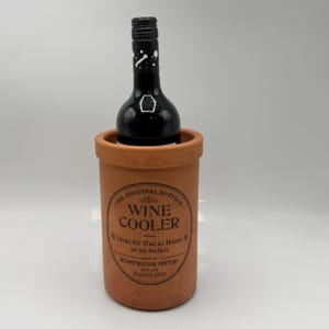 Pode incluir: Um balde de vinho de terracota com uma garrafa de vinho escuro dentro. O balde tem um rótulo com o texto "WINE COOLER" e "HENRY WATSON POTTERY". A garrafa tem uma tampa preta e um design branco.