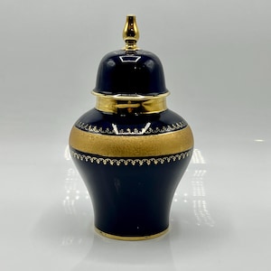 Puede incluir: Un jarrón de cerámica azul marino con una banda y detalles dorados. El jarrón tiene una tapa con un remate dorado. La banda dorada está decorada con un delicado patrón. Es un objeto decorativo.