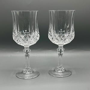 May include: A pair of clear crystal wine glasses. Each glass has a diamond-cut pattern on the bowl and a decorative stem. The glasses have a classic design, suitable for formal occasions or elegant dining.