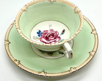 May include: A vintage teacup and saucer set with a delicate floral design. The cup and saucer are both a pale green color with a gold trim. The floral design features a pink rose with green leaves and blue flowers.