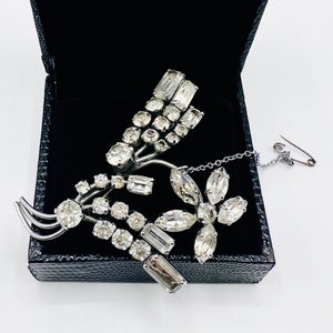 Op de afbeelding: Een zilverkleurige broche met een bloemmotief met heldere strass steentjes. De broche heeft een ketting die aan de achterkant is bevestigd.