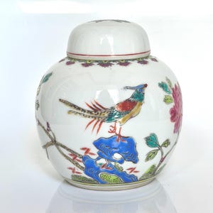 May include: White ceramic jar with lid, featuring a colorful bird perched on a blue rock, surrounded by floral and branch designs. The jar has a round shape and is decorated with red, green, and pink accents.