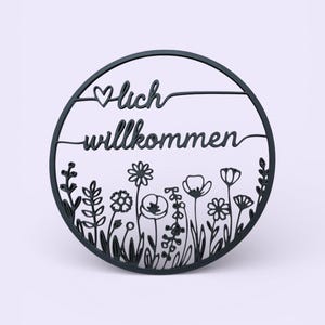Könnte beinhalten: Schwarze, kreisförmige Wanddekoration mit dem deutschen Ausdruck "lich willkommen" und einem Herzsymbol. Das Design zeigt einen floralen Rand mit verschiedenen Blumen- und Pflanzensilhouetten. Das Schild besteht aus dunklem Material und ist vor einem hellen Hintergrund platziert.
