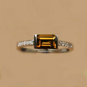 May include: A silver-toned ring featuring a rectangular, emerald-cut amber-colored gemstone set in the center. The band is accented with small, clear pavé-set stones along the sides. The text TheJewelryArts is visible on the ring band.