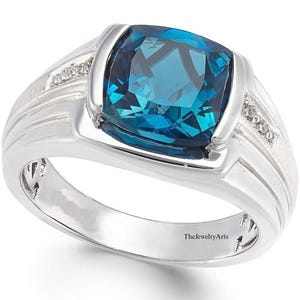 May include: A silver ring featuring a large, square-cut, teal gemstone. The ring has a wide band with etched details and small accent stones on each side of the main stone. The text "TheJewelryArts" is visible on the band.