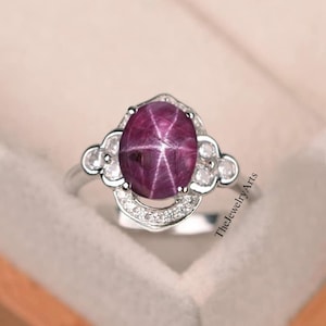 May include: A silver ring with a large, oval-shaped, red star ruby gemstone surrounded by smaller white gemstones. The ring has a delicate, ornate design.