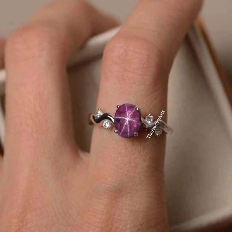 K&ouml;nnte beinhalten: Ein silberner Ring mit einem rosafarbenen Stern-Saphir-Edelstein. Der Ring hat eine zarte, wellenf&ouml;rmige Band mit kleinen wei&szlig;en Diamanten auf beiden Seiten des Edelsteins.