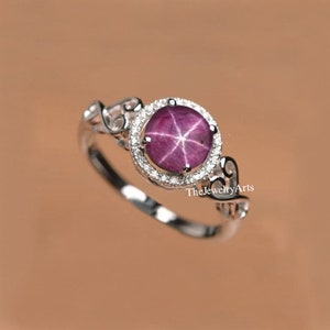 Puede incluir: Un anillo de plata con una piedra preciosa de rubí estrella rosa rodeada por un halo de diamantes más pequeños. El anillo tiene un diseño delicado y ornamentado con un patrón de espiral en la banda.