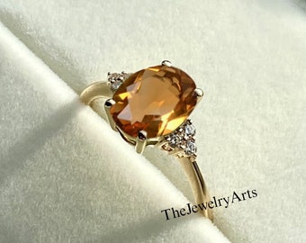 Puede incluir: Un anillo de oro con una gran piedra preciosa de corte ovalado color ámbar en el centro, flanqueada por tres pequeñas piedras claras a cada lado. La banda es fina y metálica. El texto TheJewelryArts es visible cerca de la base.