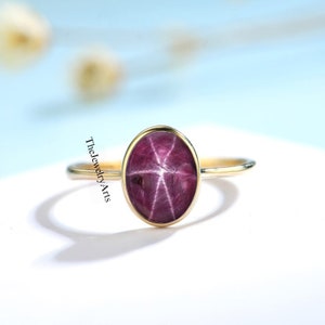 May include: A gold ring with a star ruby gemstone. The ruby is oval shaped and has a star-like pattern in the center.