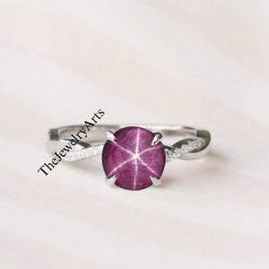 May include: A silver ring with a twisted band and a round, pink star ruby gemstone. The ring is set with small diamonds around the band.