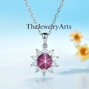 Könnte beinhalten: Eine silberne Kettenkette mit einem Anhänger, der einen rosafarbenen Sternrubin zeigt, der von einem Heiligenschein aus kleineren weißen Diamanten umgeben ist. Der Anhänger ist sonnenstrahlenförmig.