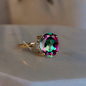 Pode incluir: Um anel de ouro com uma grande gema oval e iridescente cercada por gemas menores e transparentes. A gema apresenta tons de rosa, verde e azul.