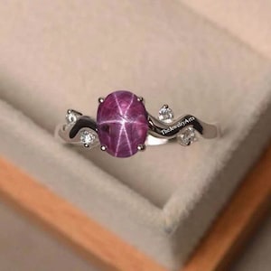 May include: A silver ring with a pink star sapphire gemstone and smaller clear gemstones. The ring has a wavy band and the text "The Jewelry Arts" is engraved on the band.