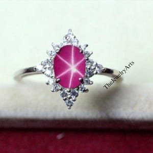 May include: A silver ring featuring a large, oval, pink star ruby gemstone. The ruby has a six-ray star effect. The ruby is surrounded by a halo of small, clear gemstones. The ring is set on a light background.