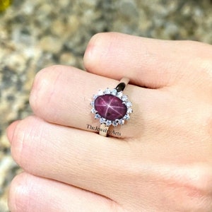 May include: A silver ring featuring a large oval ruby gemstone, surrounded by a halo of smaller white gemstones. The ruby has a star-like pattern within it.