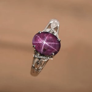 Puede incluir: Un anillo de plata con una piedra preciosa de rubí estrella. El rubí es de color rojo intenso y tiene un patrón en forma de estrella en el centro. El anillo está engastado en un delicado diseño floral.